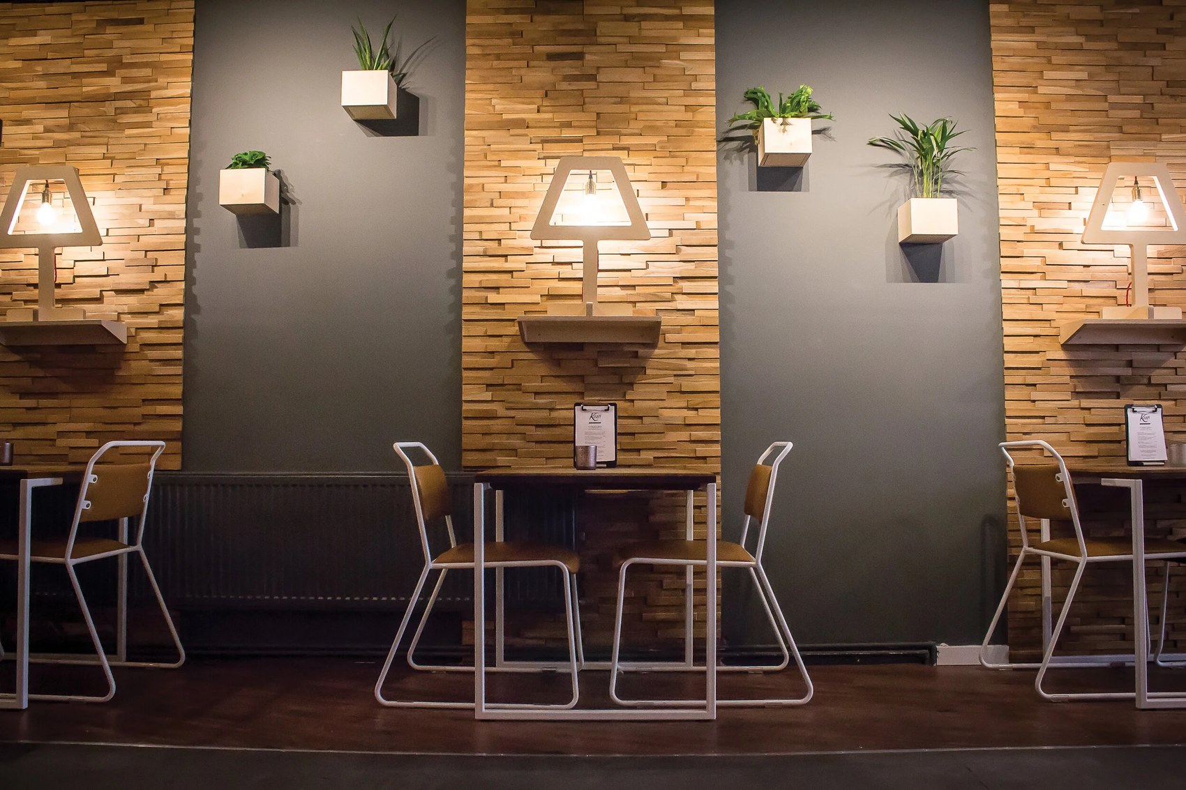 Empty tables against a wood-slat and grey wall at Kaafi, lit by small wall-mounted lamps with plants tucked between panels